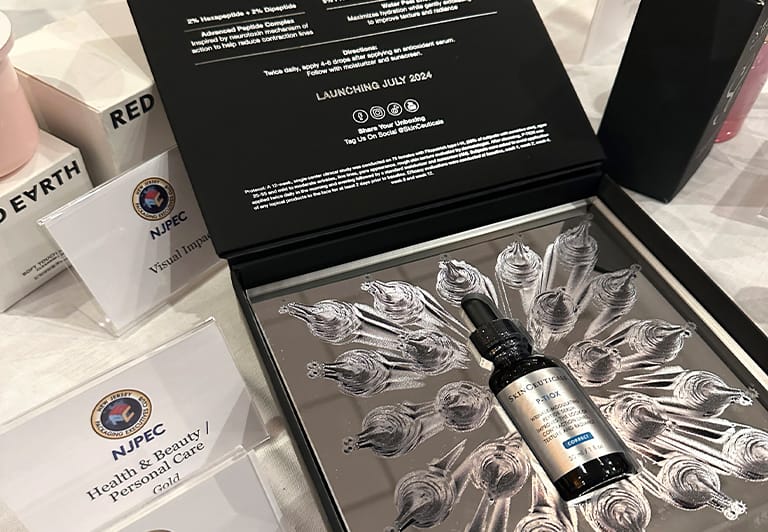 A skincare serum bottle is displayed in an open box with decorative packaging, alongside NJPEC signs for "Health & Beauty / Personal Care" and "Visual Imp.," celebrating Hatteras Brings Home Three Awards from Neographics Event.