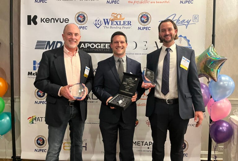 Three men in business attire stand in front of a sponsor backdrop at the Neographics Event, each holding an award and smiling at the camera. Colorful balloons are visible at the sides as Hatteras brings home three awards.