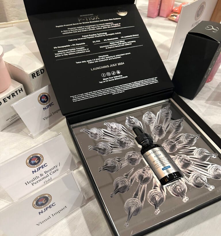 A serum bottle displayed in an open black box with a decorative interior, surrounded by NJPEC award cards and various beauty products on a table, celebrates Hatteras Brings Home Three Awards from Neographics Event.