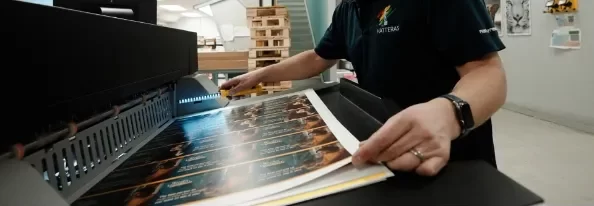 A person operates a large printer, checking a sheet of printed labels or posters emerging from the machine in an industrial workspace.