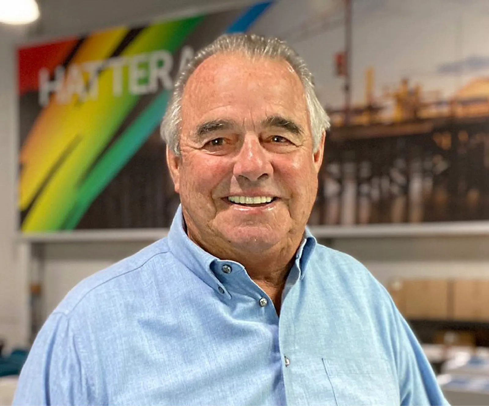 An older man with gray hair wearing a light blue button-up shirt stands indoors in front of a colorful wall mural.