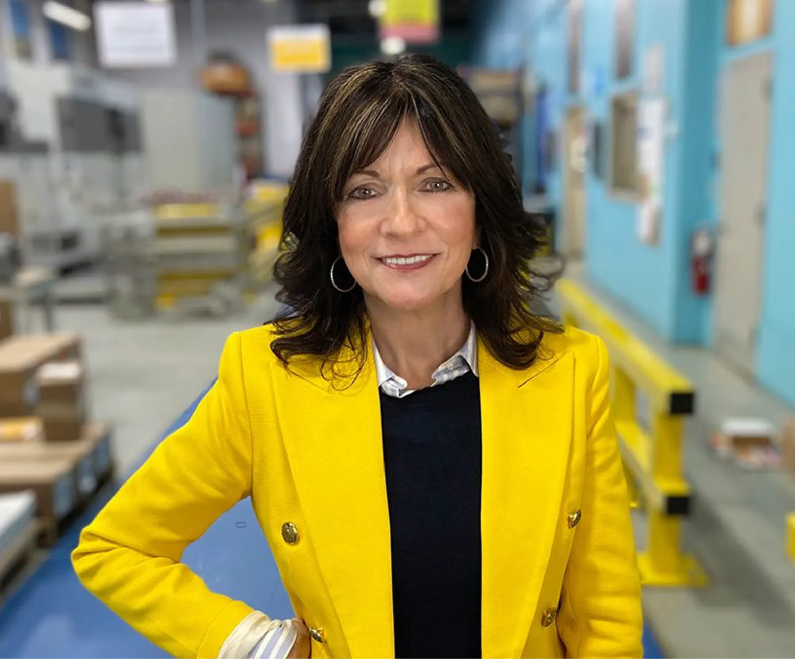 A woman wearing a yellow blazer stands indoors in an industrial or warehouse setting, smiling at the camera.