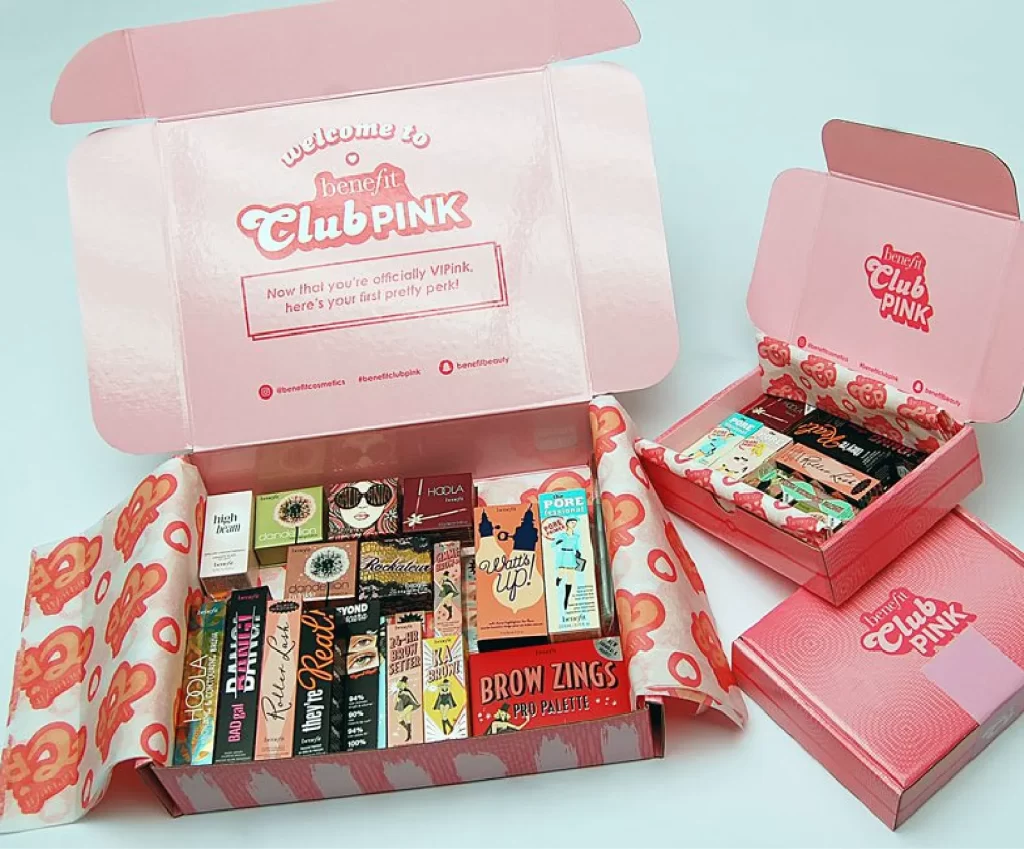 Open pink Benefit Club PINK boxes containing an assortment of Benefit Cosmetics makeup products arranged neatly inside.