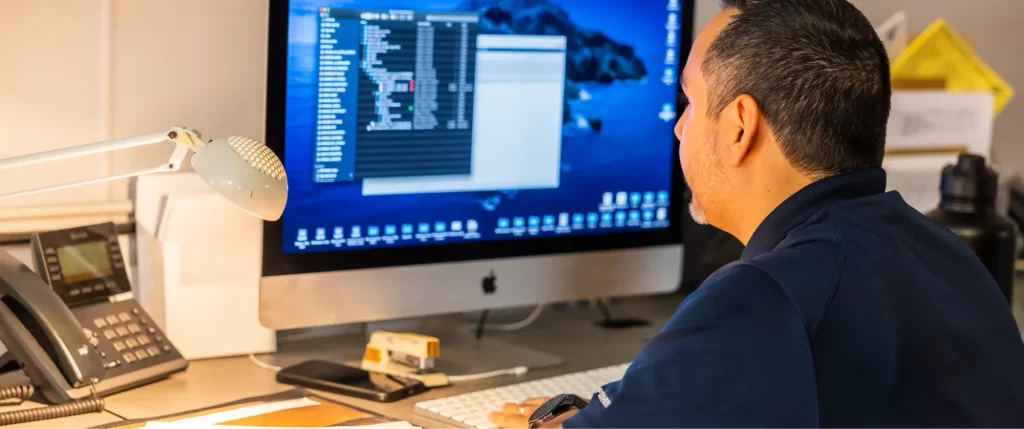 A man sits at a desk working on a computer with a large monitor displaying a file directory. Office items and a desk lamp are visible.