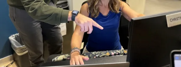 Two people at a desk look at a computer monitor; one is sitting and typing while the other stands and points at the screen.