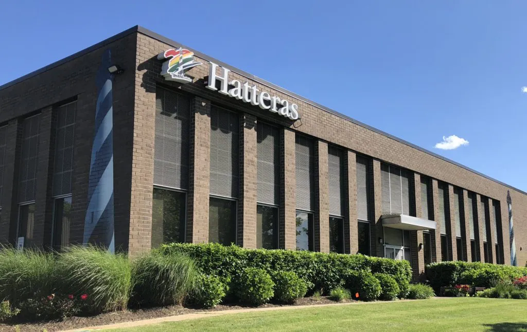 A brown brick commercial building with the "Hatteras" sign on the facade, surrounded by green shrubs and grass under a clear blue sky.