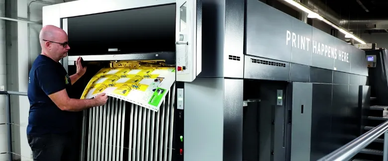 A man operates a large industrial printing machine, inspecting a sheet with multiple colorful printed images. The words "PRINT HAPPENS HERE" are visible on the machine.