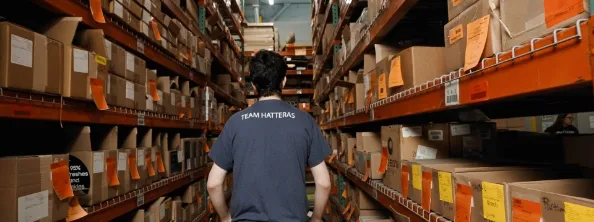 A person wearing a "Team Hatteras" shirt stands in a warehouse aisle lined with shelves filled with labeled cardboard boxes.