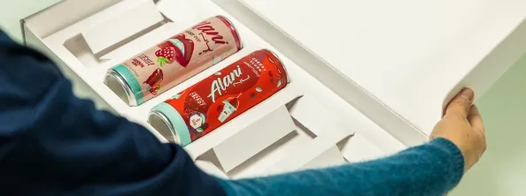 A person opens a box containing two Alani Nu energy drink cans in different flavors, each held in its own slot within the packaging.