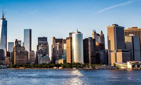 Skyline of a city with modern skyscrapers and waterfront under a clear blue sky, viewed across a body of water.