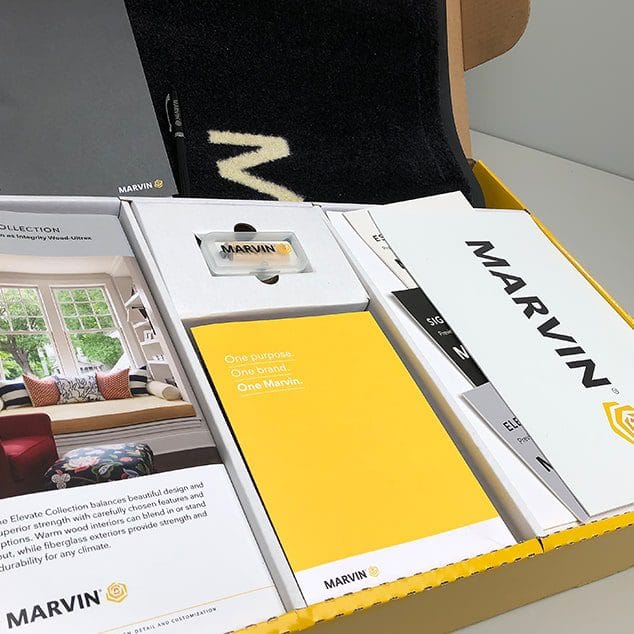 Open box containing Marvin-branded brochures, a booklet, a USB drive, and a black cloth item with a white letter "M".