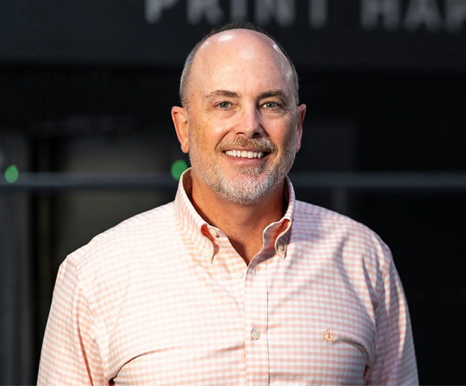 A middle-aged man with a shaved head and gray beard, wearing a light pink checkered shirt, smiling while standing indoors.