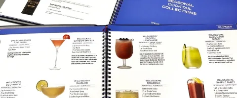 Open spiral-bound cocktail recipe book showing pages with images and descriptions of various mixed drinks, titled "Seasonal Cocktail Collections.