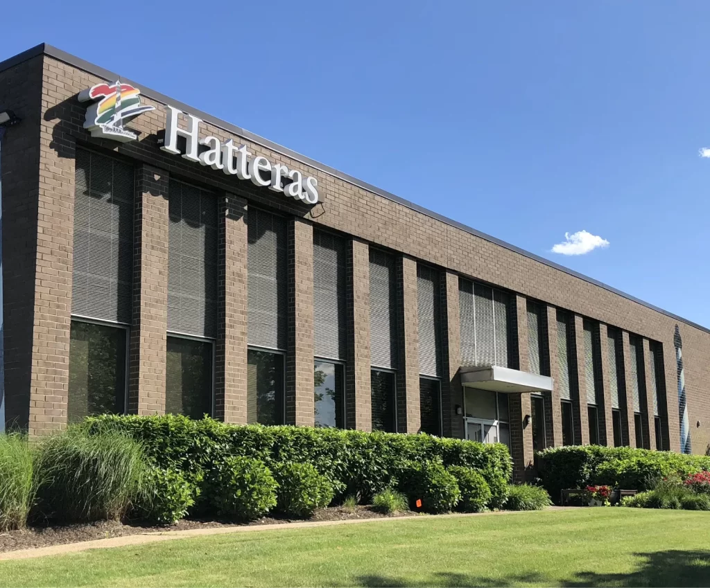A modern two-story brick building with large vertical windows and "Hatteras" signage above the entrance, surrounded by green bushes and lawn under a clear blue sky.