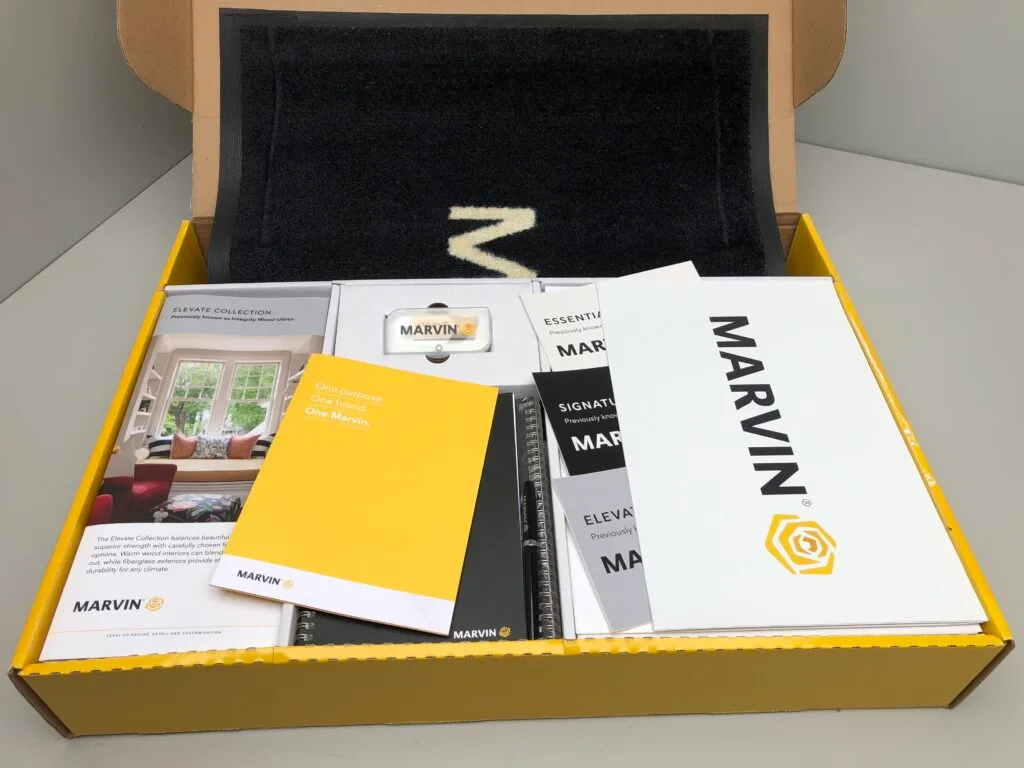 A yellow and black box contains Marvin brand brochures, booklets, a branded doormat, and marketing materials neatly arranged inside.