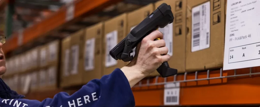 A person scans a barcode on a cardboard box in a warehouse using a handheld barcode scanner.
