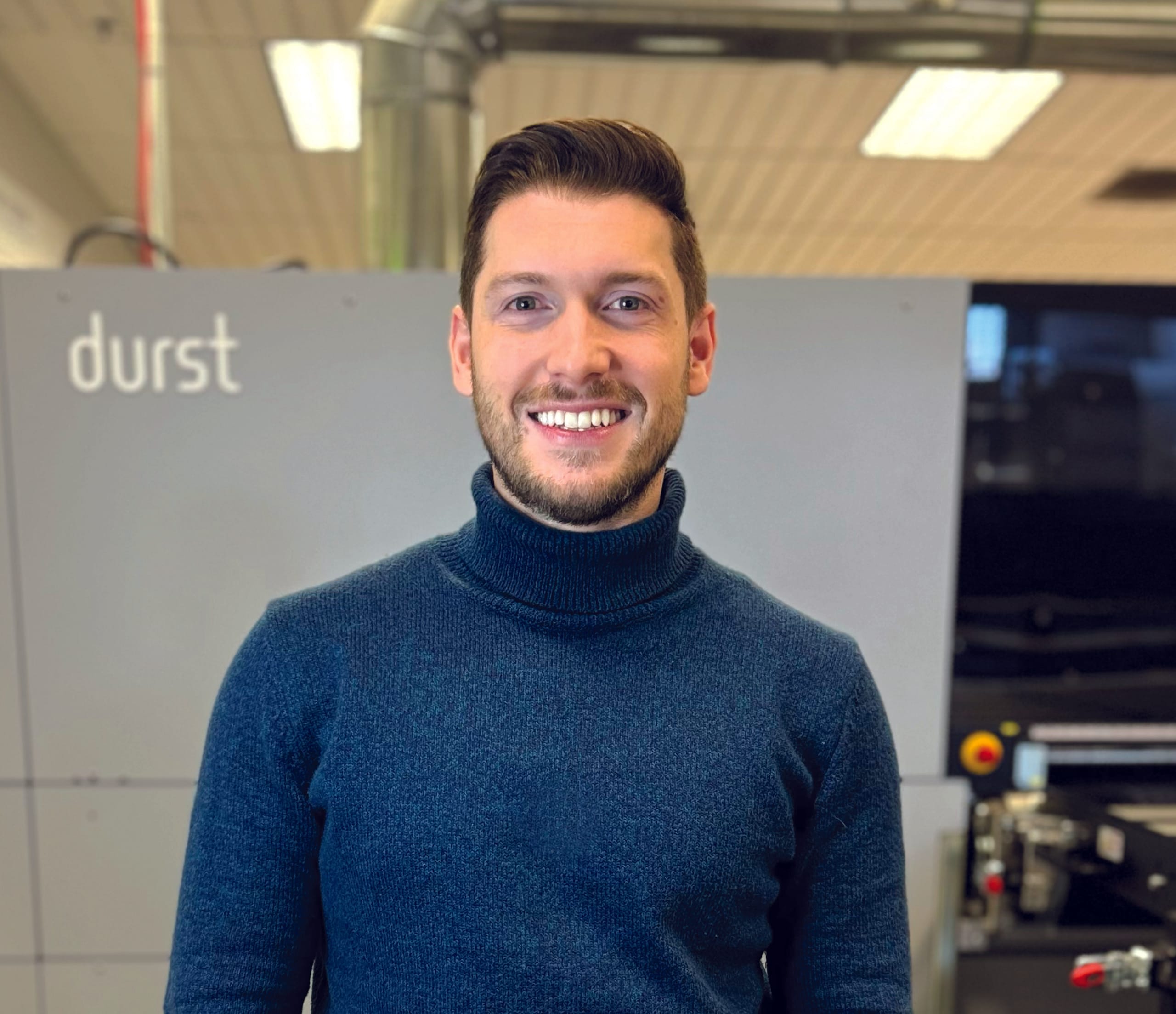 A man wearing a blue turtleneck sweater, identified as Kaue Tomasine Chaulet, stands indoors in front of industrial equipment labeled "durst," smiling at the camera.