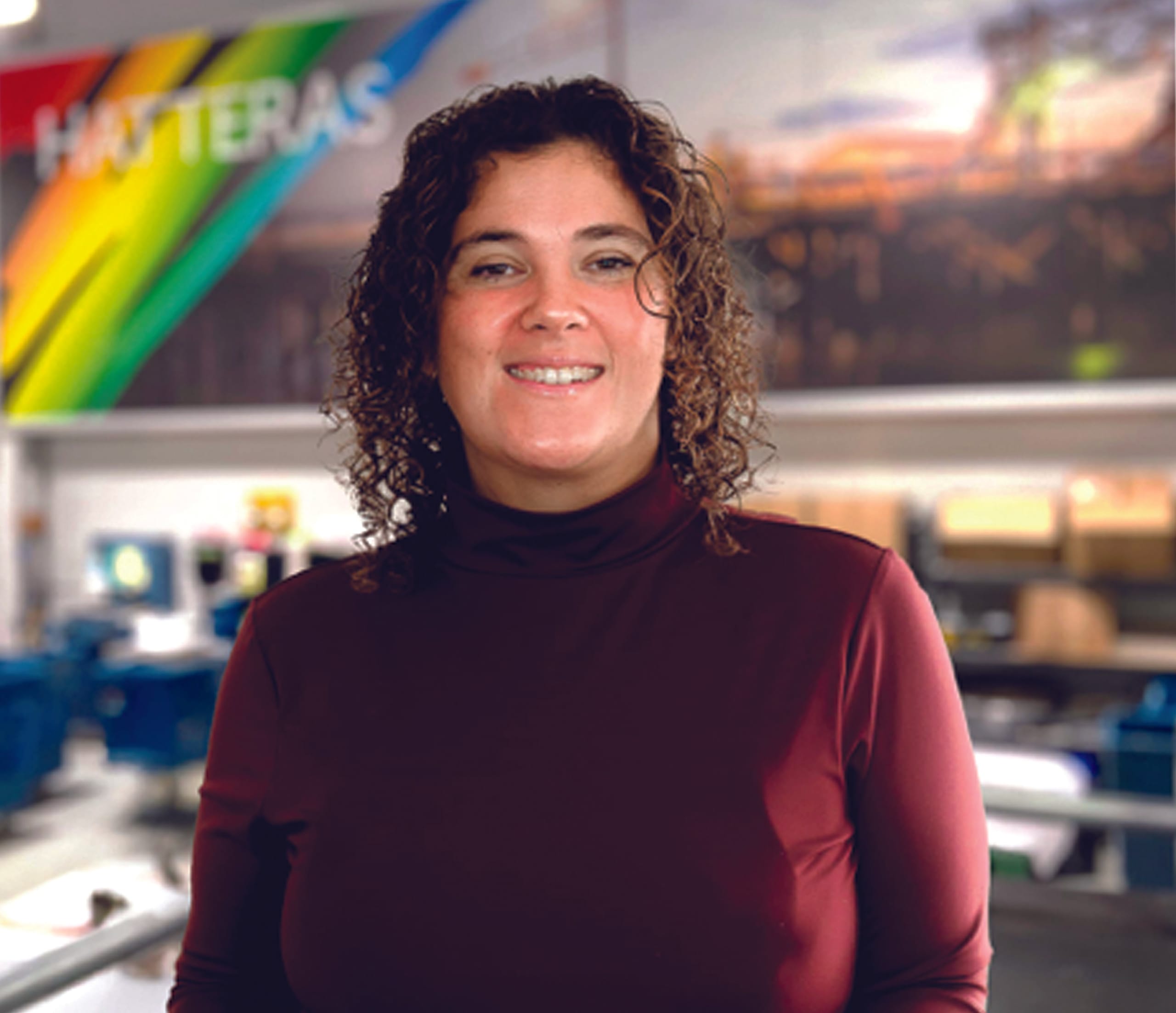 Arissa Dickison, with curly hair and a maroon turtleneck, stands indoors in front of a colorful wall sign that reads "HATTERAS.