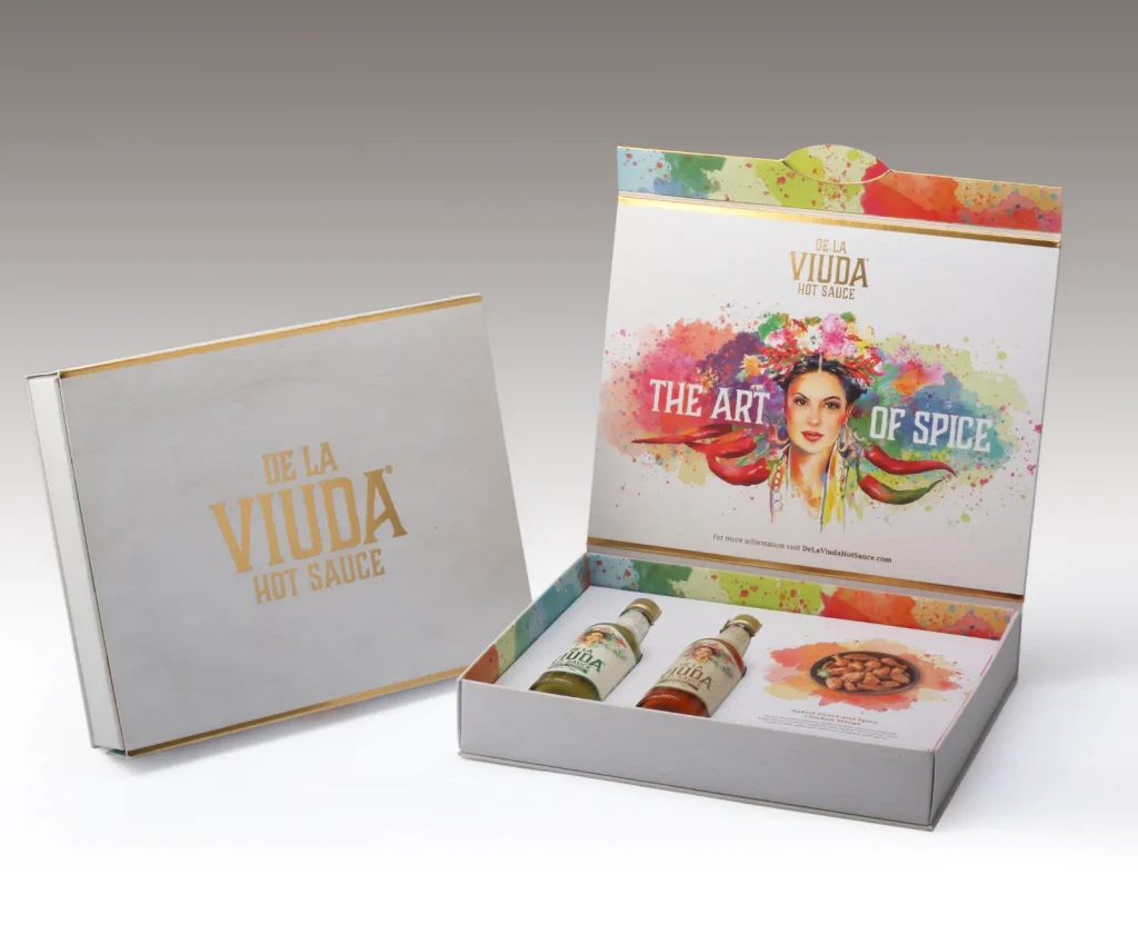 Open presentation box showcasing De La Viuda Hot Sauce set with colorful paint-splash artwork and three small sauce bottles inside. The front reads 'THE ART OF SPICE' with a vibrant Mexican-inspired design.