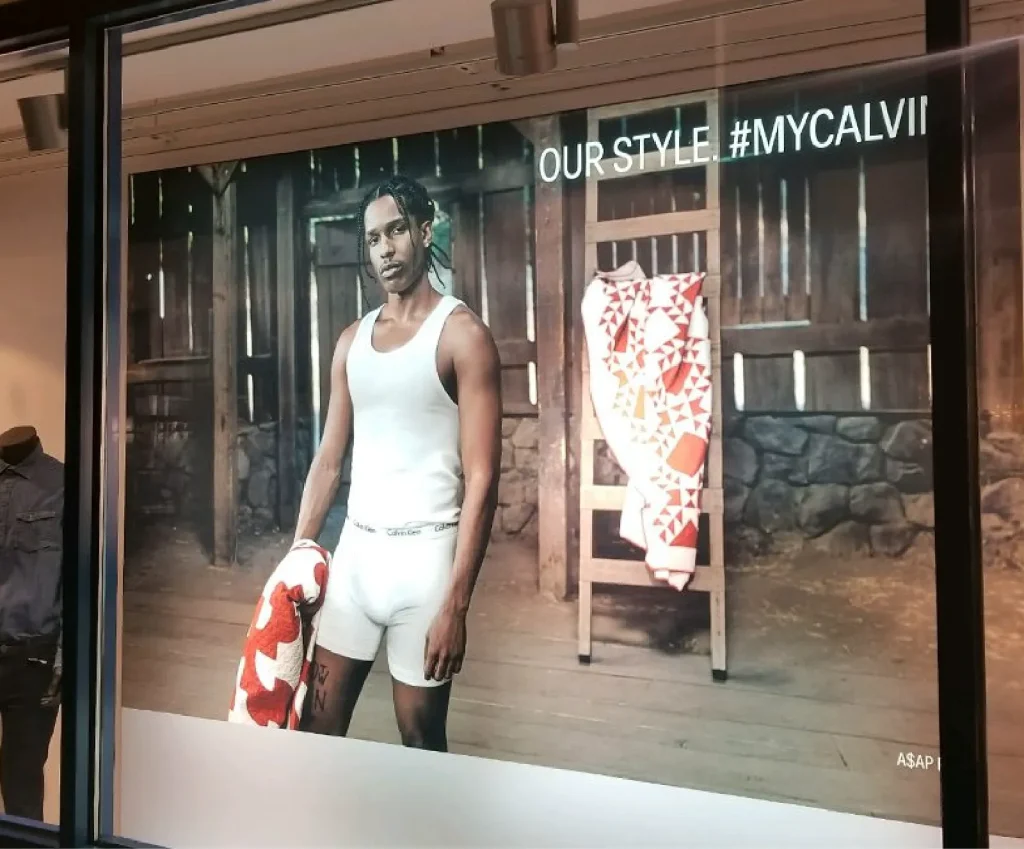 Man in white underwear holding a red-and-white towel and boxing gloves, standing in front of a rustic wooden backdrop in a store ad.