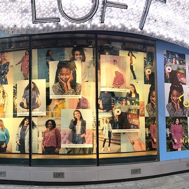 Loft storefront window display with a collage of fashion posters featuring models in colorful outfits in a curved glass storefront window area.