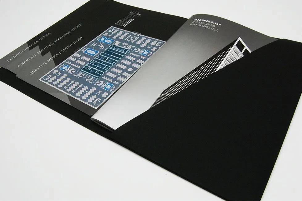 Open black portfolio with several overlapping gray brochures showcasing a blue schematic and white text, on a white surface.