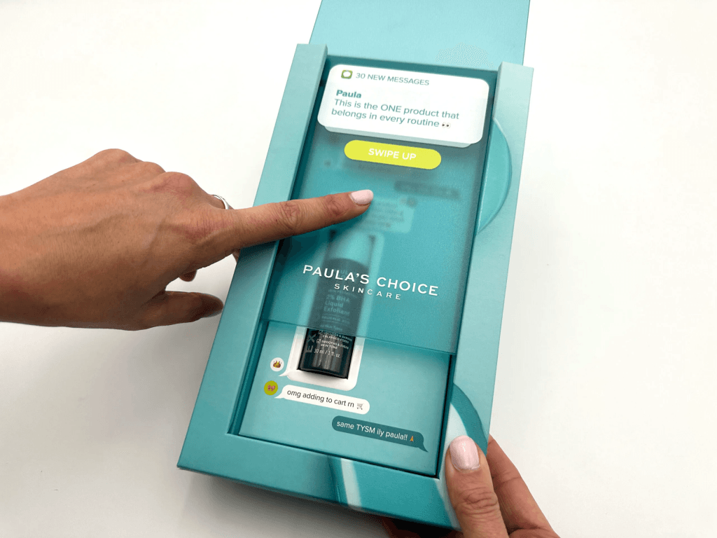 Close-up of a teal Paula's Choice skincare box with a finger tapping a yellow 'Swipe Up' button on a card inside the box