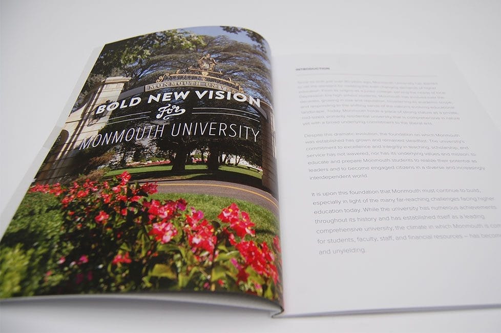 Open book showing a large photo of Monmouth University’s entrance sign with red flowers in the foreground and trees in the background.
